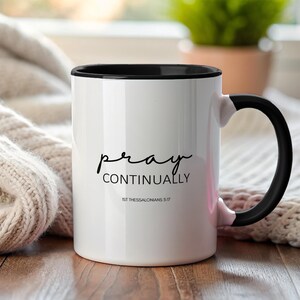 May include: White ceramic mug with a black interior and handle. The mug features the words "pray continually" in a handwritten font, along with the scripture reference 1st Thessalonians 5:17. The mug is on a wooden surface.