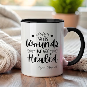 Puede incluir: Taza de cerámica blanca con interior y asa negros. La taza presenta el texto "By His Wounds We Are Healed" en escritura negra, con el versículo bíblico Isaías 53:5. La taza está sobre una superficie de madera.