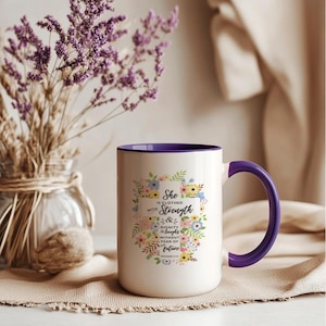 Puede incluir: Taza de cerámica blanca con interior y asa morados. La taza está decorada con un diseño floral y el texto "She is clothed with Strength & Dignity & laughs without fear of future".