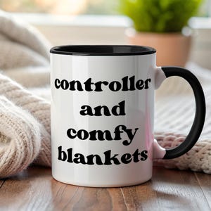 May include: White ceramic mug with black interior and handle. The mug features the words "controller and comfy blankets" in black, bold font. The mug is set against a cozy background with a knitted blanket.