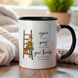 Puede incluir: Taza de cerámica blanca con interior y asa negros. La taza presenta una escalera decorativa con girasoles, una calabaza y un lazo a cuadros verdes. El texto en la taza dice: "regocijaos siempre, orad sin cesar, dad gracias en todo 1 Tesalonicenses 5:16".