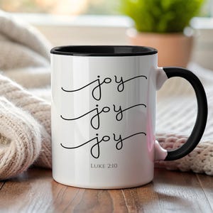 May include: White ceramic mug with a black interior and handle. The mug features the word "joy" written three times in a cursive font, with "Luke 2:10" printed below. The mug is set on a wooden surface.