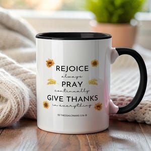 Puede incluir: Taza de cerámica blanca con interior y asa negros. La taza presenta el texto "REJOICE always PRAY continually GIVE THANKS in everything" con ilustraciones de girasoles y trigo. El texto "1ST THESSALONIANS 5:16-18" está impreso en la parte inferior.