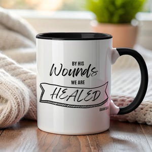 May include: White ceramic mug with a black interior and handle. The mug features the text "BY HIS Wounds WE ARE HEALED" in black script, with "Isaiah 53:5" below. The mug is set against a soft, neutral-toned background.