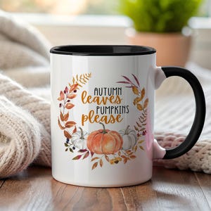 Könnte beinhalten: Weißer Keramikbecher mit schwarzem Innenraum und Henkel. Der Becher zeigt ein Aquarelldesign aus Herbstblättern und Kürbissen mit dem Text "AUTUMN leaves PUMPKINS please" in einer dekorativen Schriftart.