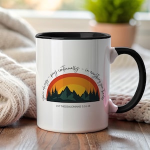 Puede incluir: Taza de cerámica blanca con interior y asa negros. La taza presenta un diseño colorido de una puesta de sol sobre montañas, con el texto "rejoice always - pray continually - in everything give thanks" y "1ST THESSALONIANS 5:16-18".