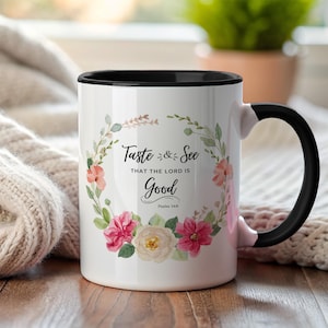 Puede incluir: Taza de cerámica blanca con interior y asa negros. La taza presenta un diseño de corona floral con el texto "Taste & See That The Lord Is Good" en escritura negra. Las flores son rosas, melocotón y blancas.