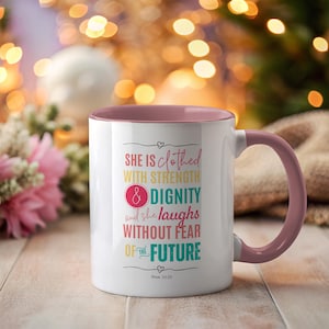 Puede incluir: Taza de cerámica blanca con interior y asa rosas. La taza presenta el texto "SHE IS clothed WITH STRENGTH & DIGNITY and she laughs WITHOUT FEAR OF THE FUTURE" en varios colores. También está impreso el versículo bíblico Proverbios 31:25.