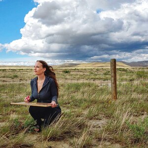 May include: A person kneels in a field, holding a wooden board and a red object. The scene is set against a backdrop of a cloudy sky and a wire fence. The person is wearing a dark blue hooded jacket and black pants.