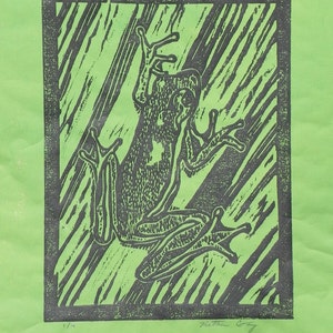 May include: A black and white linocut print of a frog on a green background. The print is titled "The Frog" and is numbered 1/4.