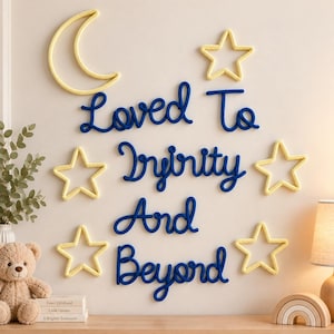Op de afbeelding: Wanddecoratie met donkerblauwe touwletters die de tekst Loved To Infinity And Beyond vormen, omringd door een gele touwmaan en vier gele touwsterren. Een pluche beer en boeken staan op een houten ondergrond.