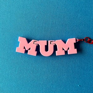 May include: A pink necklace spelling out the word "MUM" in capital letters. The letters are connected and attached to a red chain. The background is a solid teal color.