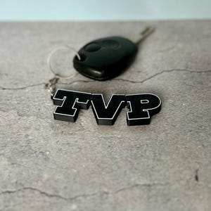 May include: Black and white TVP keychain with a car key. The keychain features the letters "TVP" in a bold, blocky font. The car key is black and has a silver key ring attached to the keychain.