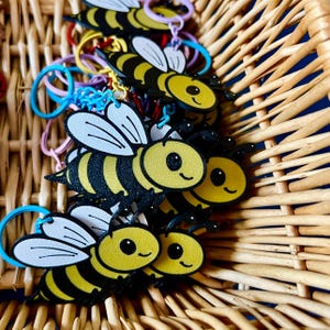 May include: A collection of bee-shaped keychains with yellow and black striped bodies, white wings, and smiling faces. The keychains are attached to colorful rubber rings and are arranged inside a woven basket.