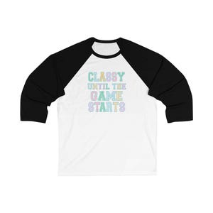 Puede incluir: Una camiseta de béisbol blanca con mangas negras. La camiseta presenta el texto "CLASSY UNTIL THE GAME STARTS" en colores pastel. La camiseta está hecha de un material suave y es adecuada para ropa casual.