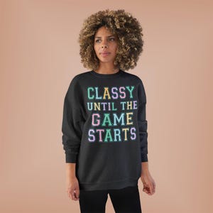 Puede incluir: Una sudadera gris oscuro con el texto "CLASSY UNTIL THE GAME STARTS" en colores pastel. La sudadera tiene cuello redondo y mangas largas. El tema general es ropa casual.