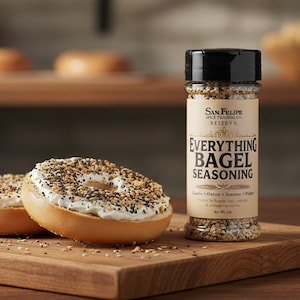 Puede incluir: Dos bagels con queso crema y condimento para bagels, sobre una tabla de madera, junto a un frasco de San Felipe Spice Trading Co. Reserva Everything Bagel Seasoning. El frasco tiene una tapa negra y una etiqueta con el nombre del producto.