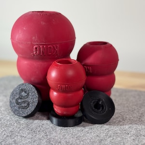 Puede incluir: Tres juguetes para perros Kong de goma roja de diferentes tamaños. El juguete más grande es un Kong clásico con el logotipo de Kong visible. Los dos juguetes más pequeños son Kongs con una abertura más pequeña. Los juguetes están sobre una superficie gris.