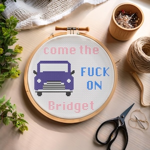 Puede incluir: Un bastidor de punto de cruz con un diseño de coche morado y el texto "come the FUCK ON Bridget" en rosa y azul. El bastidor está sobre una superficie clara con materiales de artesanía, incluyendo tijeras y un alfiletero de madera.