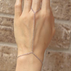 May include: A silver hand chain, also known as a hand harness, with fine chain links. The chain connects to a ring and wraps around the wrist, creating a stylish accessory. The jewellery is displayed against a neutral background.