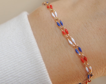 May include: A gold-toned bracelet featuring a chain of alternating red, white, and blue enamel oval links. The bracelet is displayed on a wrist, with a white long-sleeved shirt in the background. The bracelet's design is reminiscent of a flag.