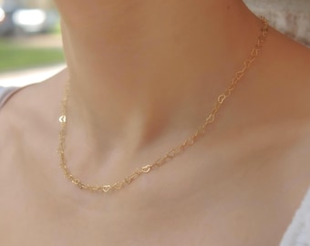 May include: A delicate gold necklace featuring a repeating heart design. The necklace is draped around the neck, showcasing the intricate heart-shaped links. The warm gold color contrasts with the skin tone, creating a subtle, elegant accessory.