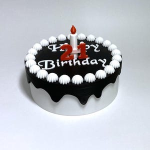 Pode incluir: Bolo redondo preto e branco com a inscrição "Happy 21 Birthday". Decorado com glacê branco e encimado por uma vela com chama vermelha e o número "21" em vermelho.