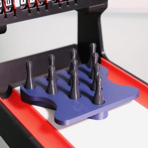 May include: A tabletop skee-ball game with a black frame, red lanes, and a blue pin platform. The game includes gray skee-balls and a scoring mechanism at the top. The game is designed for indoor entertainment.