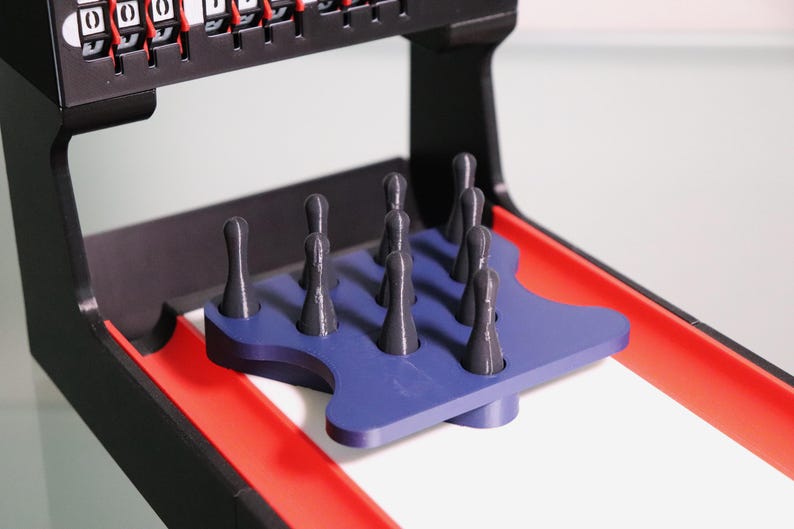 May include: A tabletop skee-ball game with a black frame, red lanes, and a blue pin platform. The game includes gray skee-balls and a scoring mechanism at the top. The game is designed for indoor entertainment.