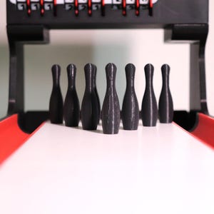 May include: A miniature bowling game set with seven black bowling pins standing on a white lane. The lane has red side rails, and a black frame with a scoring mechanism is visible in the background.