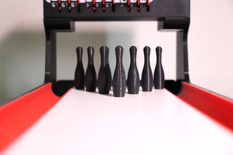 May include: A miniature bowling game set with seven black bowling pins standing on a white lane. The lane has red side rails, and a black frame with a scoring mechanism is visible in the background.