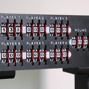 May include: A black game scorekeeper with white and red number dials. The scorekeeper has six player slots and a round counter. The text "PLAYER" and numbers 1-6 are printed above the player slots.