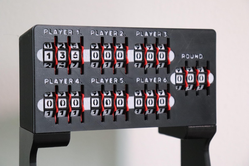 May include: A black game scorekeeper with white and red number dials. The scorekeeper has six player slots and a round counter. The text "PLAYER" and numbers 1-6 are printed above the player slots.
