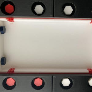 May include: A rectangular, modular construction set with a white interior and black exterior. The set features red, white, and blue accents, including hexagonal and circular elements, and red clips. The design is geometric and modern.