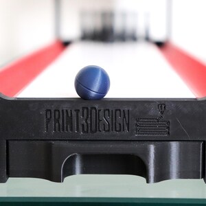 May include: A miniature skee-ball game with a black base and red side rails. A blue ball rests on the front edge, above the text "PRINT3DESIGN". The game is set on a glass surface.