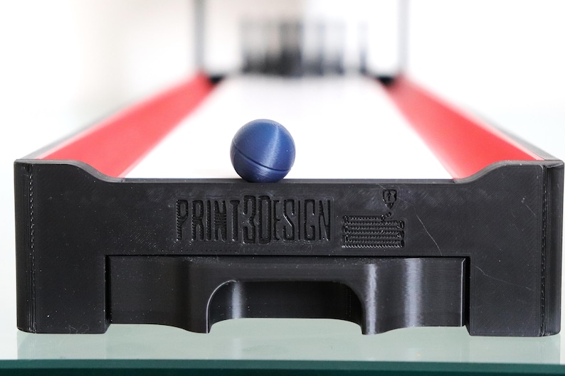 May include: A miniature skee-ball game with a black base and red side rails. A blue ball rests on the front edge, above the text "PRINT3DESIGN". The game is set on a glass surface.