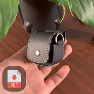 May include: A black leather case with a metal ring and a snap closure. The case is designed to hold small items, such as earbuds or a phone charger.