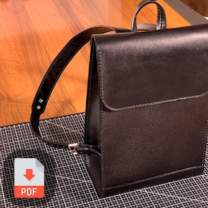 Peut inclure: Un sac à dos en cuir noir avec une seule sangle et une fermeture à rabat. Le sac à dos est posé sur une surface à carreaux noirs et blancs.
