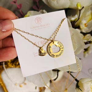 May include: A gold-colored necklace set featuring two pendants. One pendant is a heart with the word "Amor", and the other is a circle with the words "Amor de mama". The necklace is displayed on a white card with the brand name "Lorena Bracho".