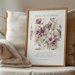 Puede incluir: Un estampado enmarcado con un diseño floral en acuarela. El estampado presenta flores moradas y rosas, hojas verdes y el texto "Rose of Sharon". El marco es de madera marrón claro. El estampado descansa sobre una silla beige con una manta.