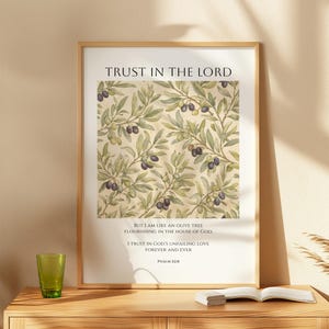 May include: A framed print featuring the text "TRUST IN THE LORD" and an olive branch design with olives. The print also includes a Bible verse. The frame is light brown, and the print is displayed on a wooden cabinet.
