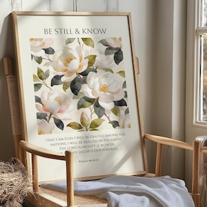 Può includere: Una stampa artistica incorniciata con un design floreale ad acquerello di fiori di magnolia bianchi e rosa e foglie verdi. Il testo "BE STILL & KNOW" è in alto, con un versetto biblico sotto. L'opera d'arte è esposta su una sedia di legno.