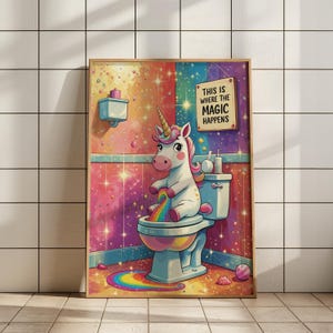 May include: A framed print depicting a unicorn on a toilet, with a rainbow. The bathroom scene is colourful with pink, blue, and gold tones, and the text reads "THIS IS WHERE THE MAGIC HAPPENS". The print is set against a tiled wall.