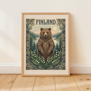 May include: Framed art print depicting a brown bear with patterned fur, set against a lake and forest. The word "FINLAND" is displayed at the top. The artwork is framed in a light wood.