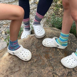 Pu&ograve; includere: Diverse paia di zoccoli bianchi con calzini a fantasia colorata. I calzini presentano vari colori, tra cui blu, verde, rosa e grigio. Gli zoccoli sono su una grande roccia vicino a un ruscello. Gli zoccoli hanno un cinturino e fori di ventilazione.