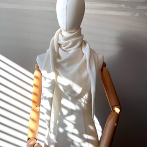 以下が含まれることがあります： マネキンの上にドレープされたクリーム色のショール。 ショールは柔らかいニット素材で作られており、首と肩の周りにレイヤー効果を生み出すようにスタイリングされています。 マネキンは、ブラインドを通して太陽光が差し込む壁の前に配置されています。