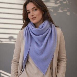 以下が含まれることがあります： 女性が、水色の三角スカーフを着用しています。スカーフは首と肩にかけられています。ベージュのセーターと、お揃いのカーディガンを着ています。背景は、影のあるニュートラルなグレーです。