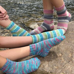 Pu&ograve; includere: Primo piano di diversi paia di calzini lavorati a mano. I calzini presentano motivi a righe in blu, rosa, verde e grigio. I calzini sono indossati da persone sedute vicino a un corso d'acqua.