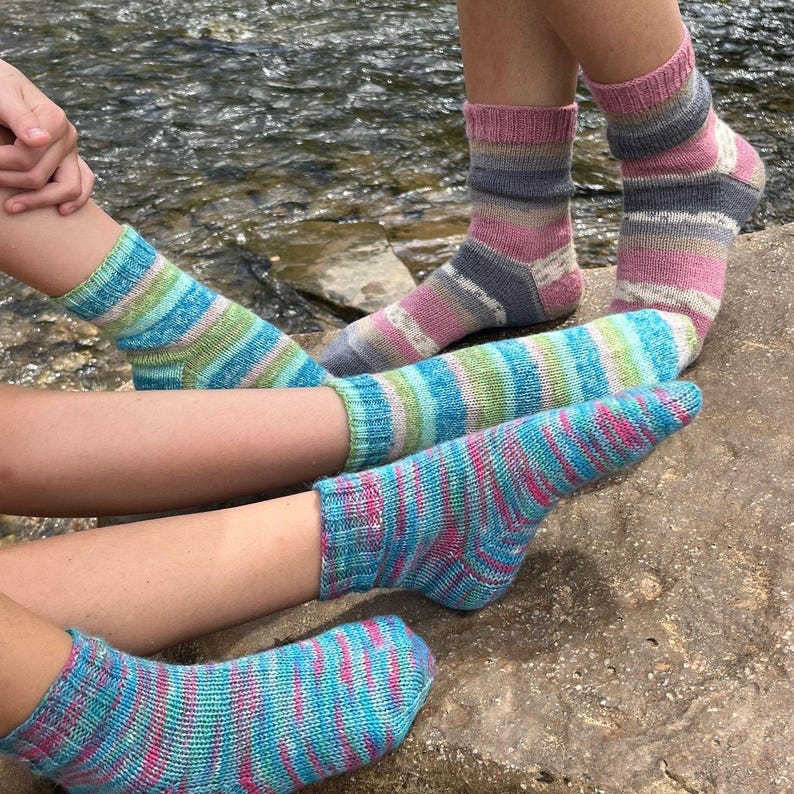 Pu&ograve; includere: Primo piano di diversi paia di calzini lavorati a mano. I calzini presentano motivi a righe in blu, rosa, verde e grigio. I calzini sono indossati da persone sedute vicino a un corso d'acqua.