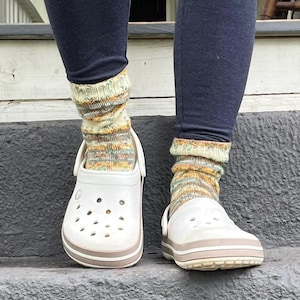 Pu&ograve; includere: Zoccoli bianchi con finiture beige indossati con calzini a maglia colorati. I calzini hanno un mix di tonalit&agrave; gialle, verdi e arancioni. La persona indossa leggings blu scuro. Gli zoccoli sono su una superficie grigia.